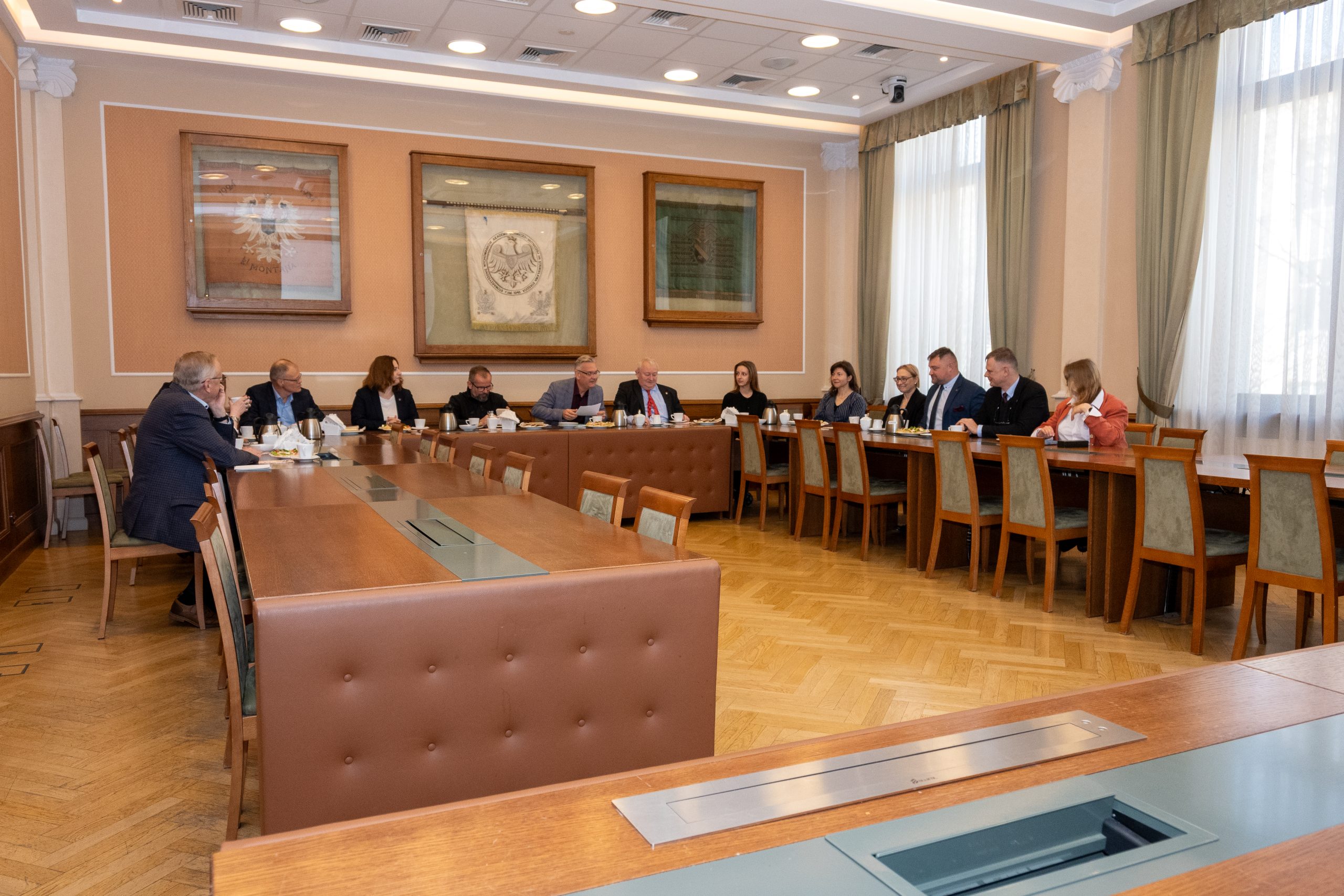Pomieszczenie konferencyjne, trzynastu gości siedzi przy brązowym stole, który ma kształt litery U. Na ścianie wiszą trzy ramy w których znajdują się herby AGH, po prawej stronie są dwa okna. Jest dzień.